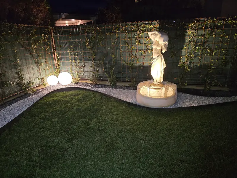 Giardino illuminato di notte con prato, bordo in ghiaia bianca e statua